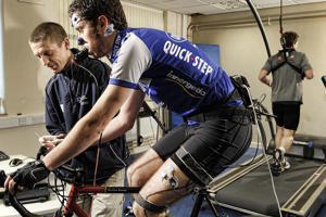 A sports analyst takes scientific readings from a man on a stationary bicycle. The man on the bicycle has measuring equipment strapped to his face.
