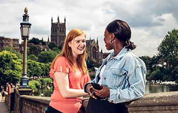 2 students stood smiling and facing each other on Worcester bridge, with Worcester Cathedral in the background