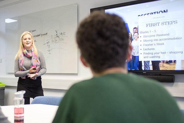 A blonde girl is giving a presentation to two students