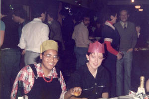 Two women in 1980s clothing and paper party hats sit in front of party food