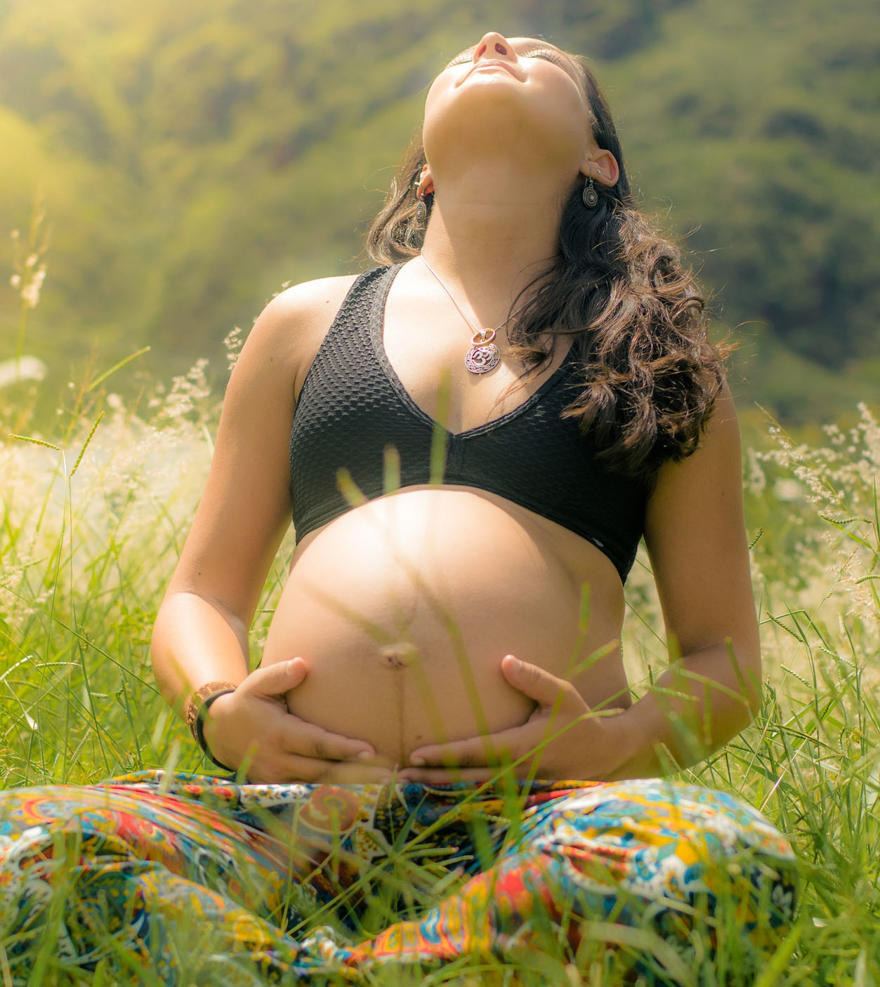 A pregnant lady is holding her bump with both hands and sitting in a field.